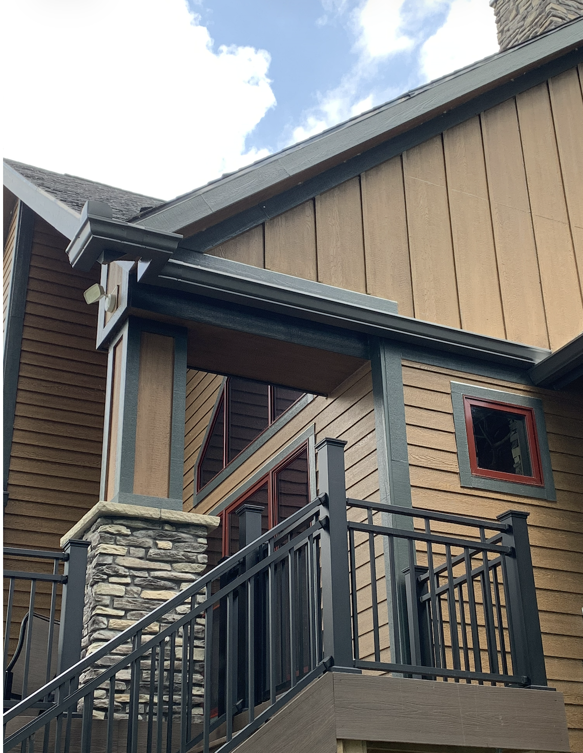Close-up of a modern house exterior featuring wooden siding, stone pillar, metal railing, and small windows, with blue sky and clouds in the background.