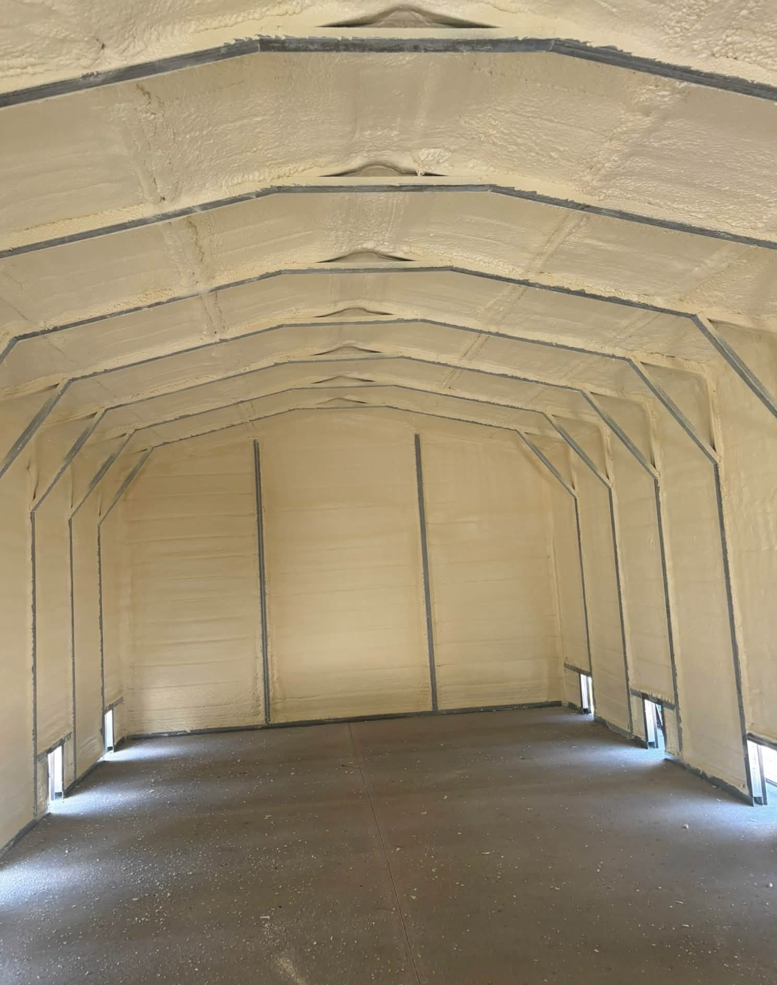 Interior of a partially insulated garage or storage space with spray foam insulation on walls and ceiling, exposed metal framing, and small windows near the floor.