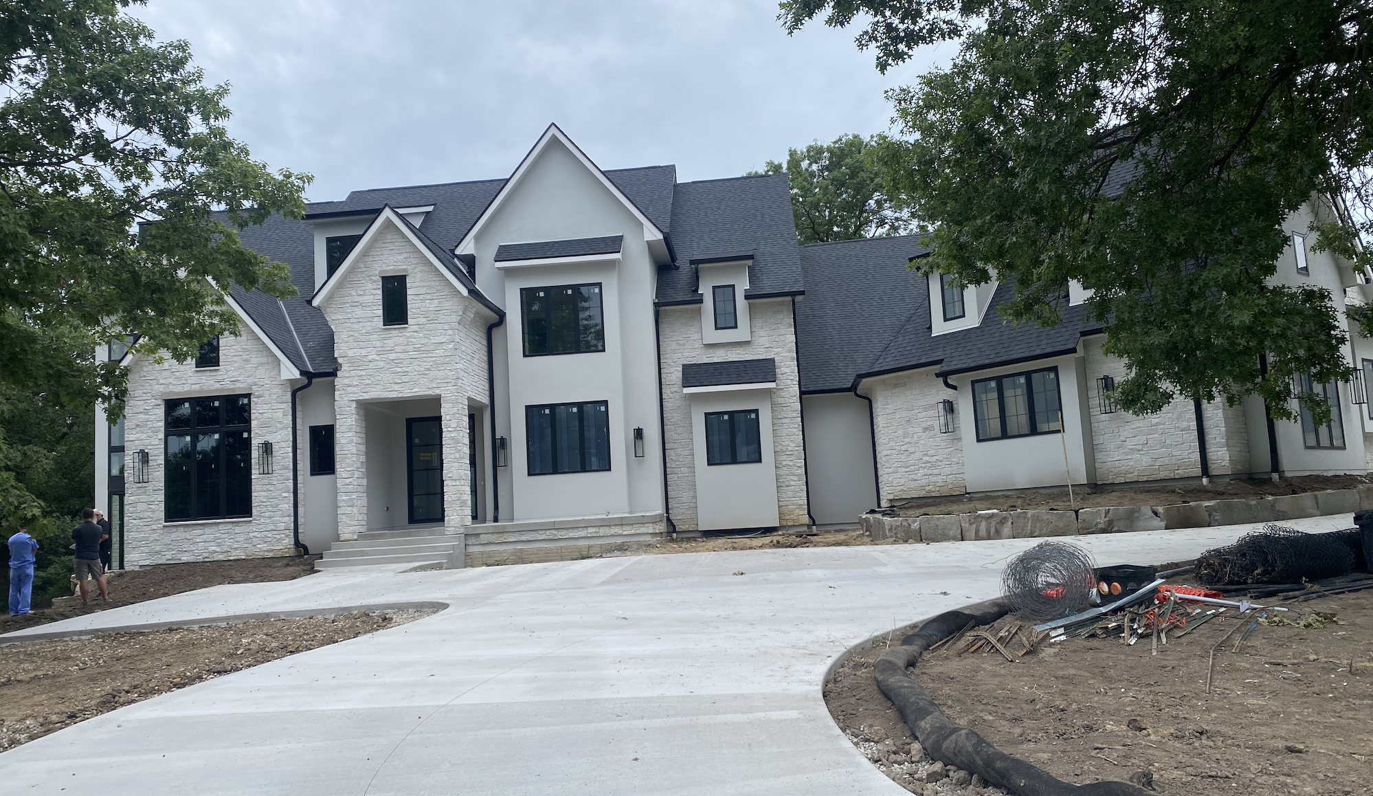Newly constructed modern multi-story house with white stone and stucco exterior, black window frames, and dark gray roof, surrounded by trees and construction materials.