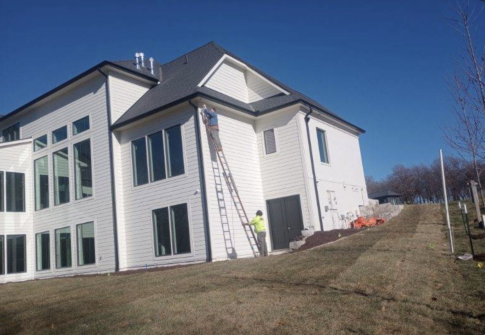 Construction workers working on the exterior of a large, white modern house with multiple windows, situated on a grassy hill on a clear sunny day.