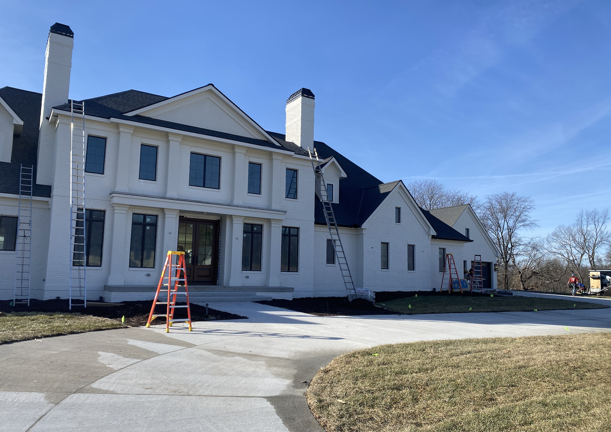 Front view of a large white house under construction with ladders and construction equipment outside. The house has multiple windows, two chimneys, and a black roof. It's sunny with clear skies, and some workers are visible in the yard.