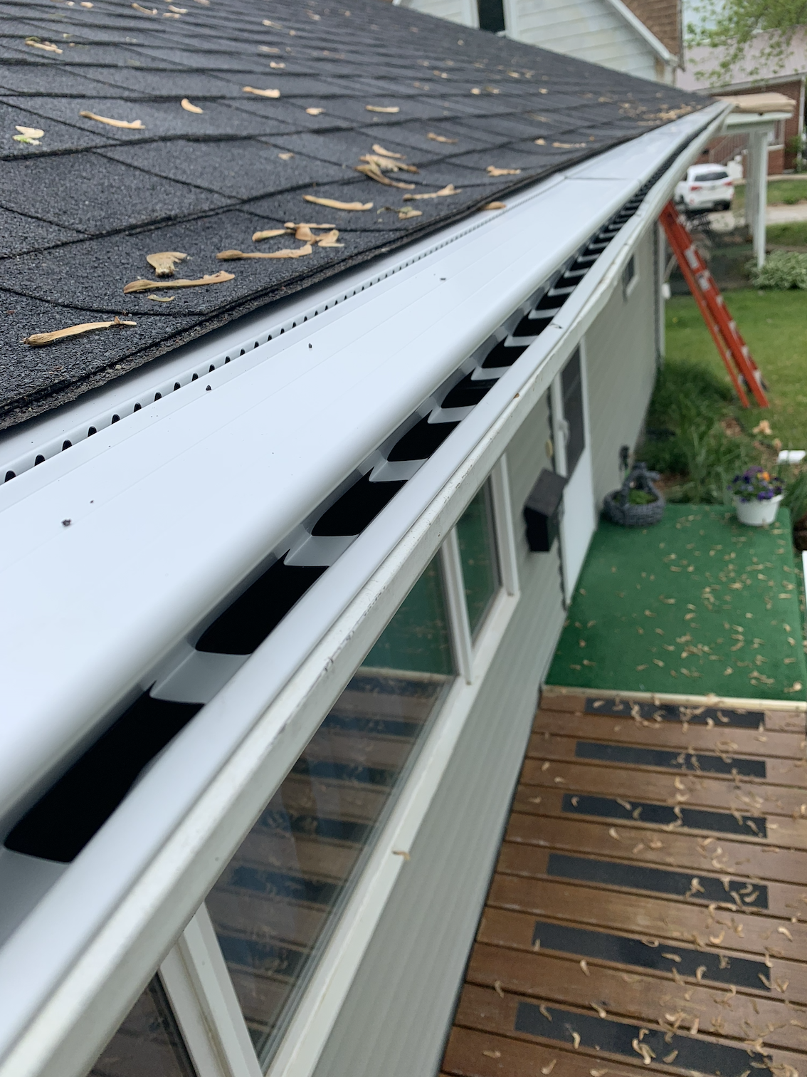 Close-up of a house's gutter and roof with fallen leaves, part of the house's backyard with a wooden deck, potted plants, and an extension ladder.