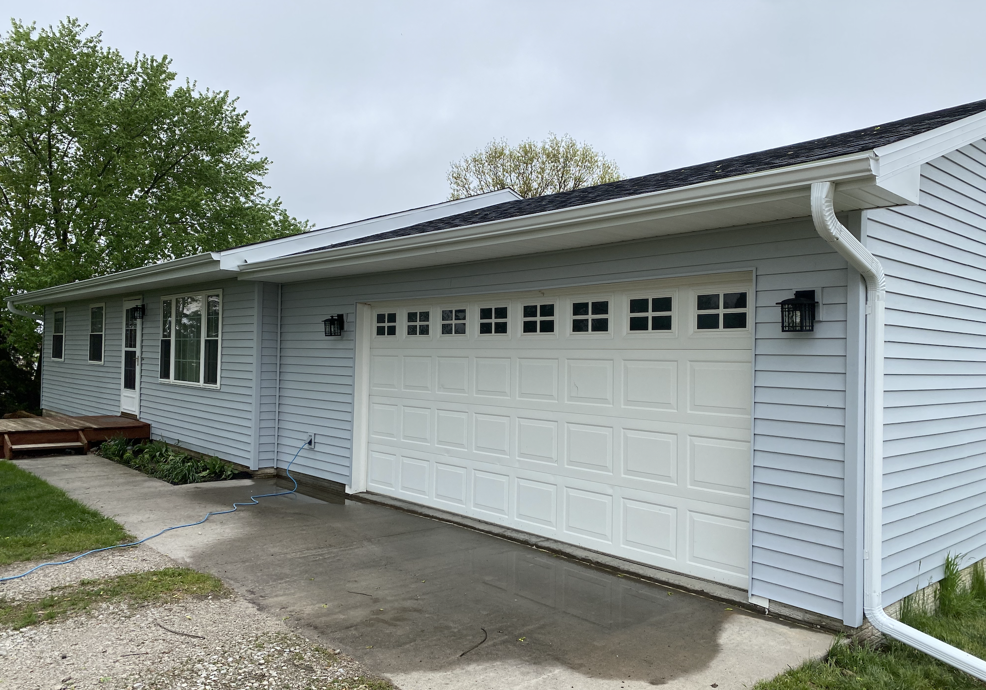 A suburban house with white vinyl siding, a white garage door, and three black outdoor wall-mounted lantern lights. There is a concrete driveway and a small garden bed with green plants and a wooden platform on the left. Two large trees with green le