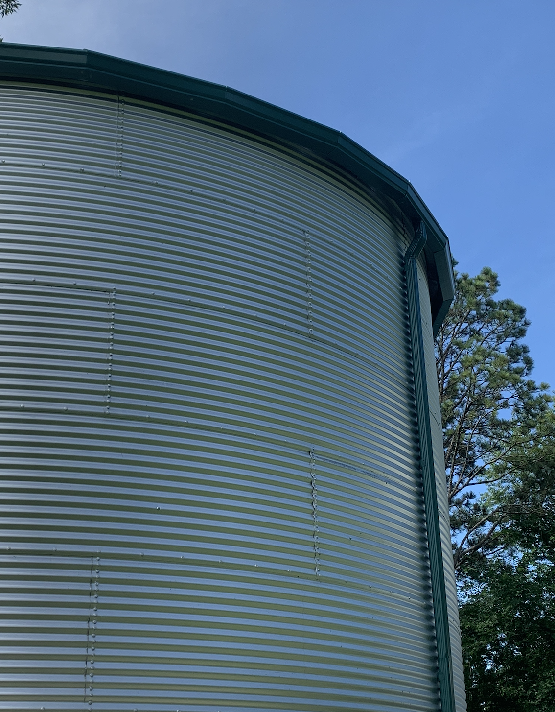 Close-up of a large, cylindrical, metal grain silo with a green roof and exterior pipe, against a clear blue sky with green trees in the background.