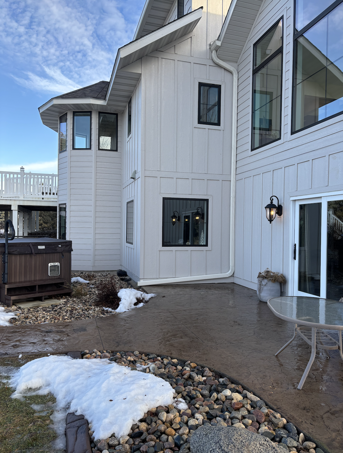 Backyard patio with a round glass table, hot tub, snow patches, and a two-story white house with multiple windows and exterior lighting against a blue sky with clouds.