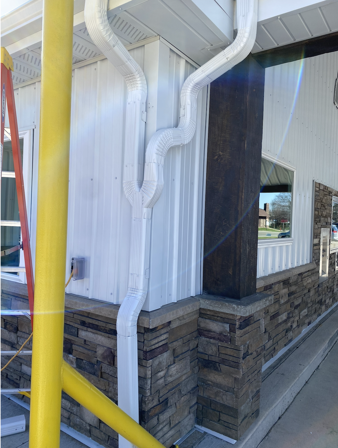 Exterior of a building with white ribbed metal siding, a stone foundation, and a large dark wooden post. White insulated flexible ducts are installed on the wall. A yellow ladder and a yellow pole are visible in the foreground.