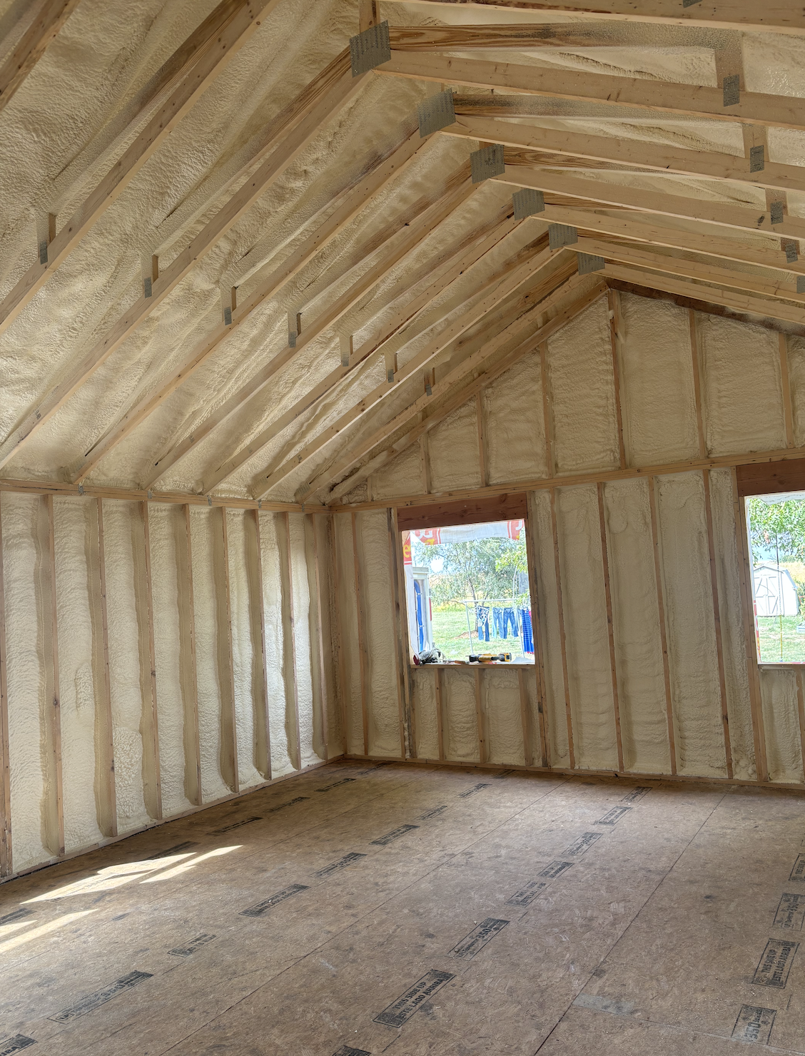 Interior view of a house under construction showing framing, spray foam insulation, and plywood subflooring, with windows opening to the outdoors.