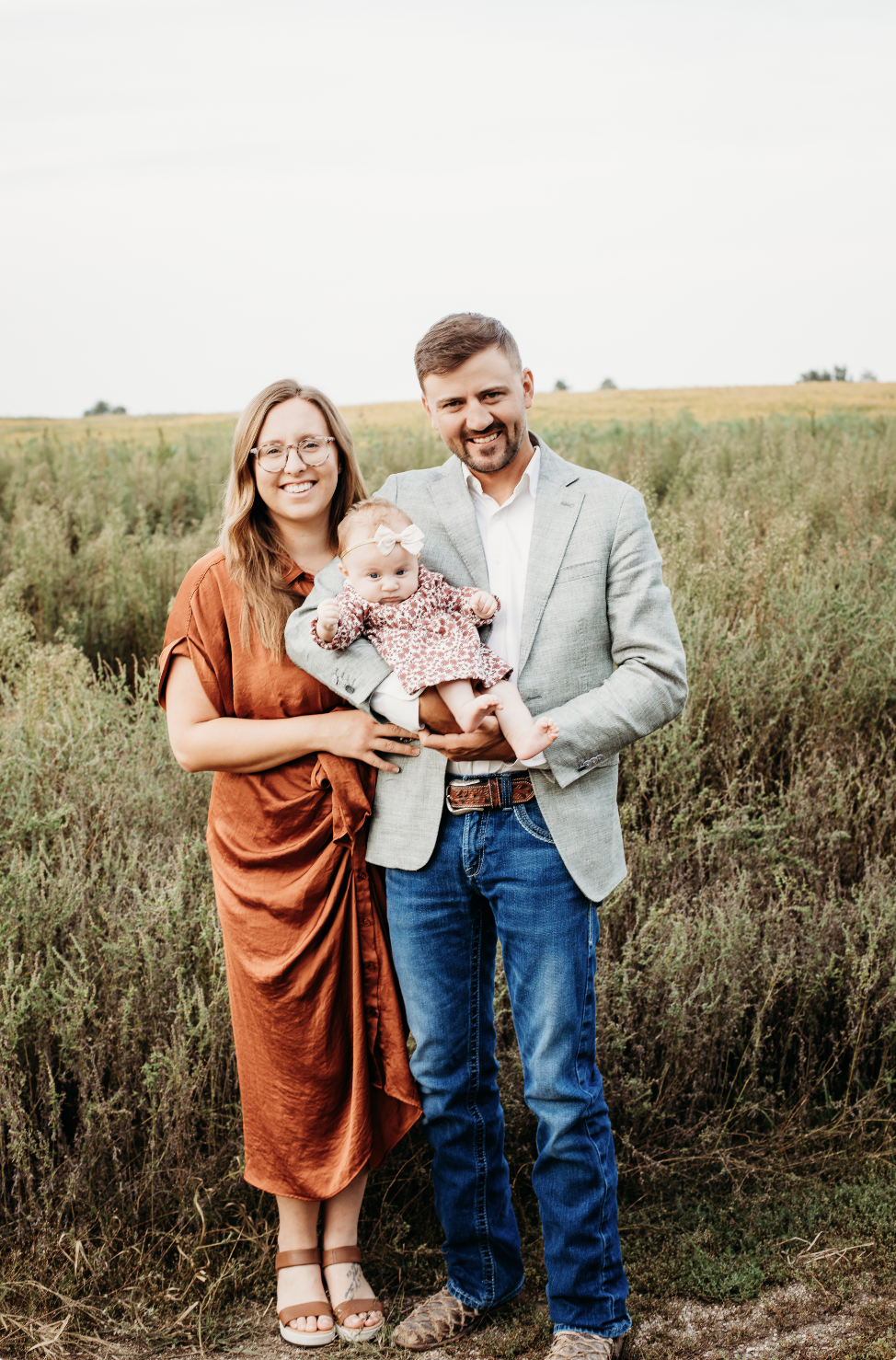 A family of three standing in a field on a cloudy day, with a woman in a brown silk dress, a man in a gray blazer and jeans, and a baby girl in a floral dress.