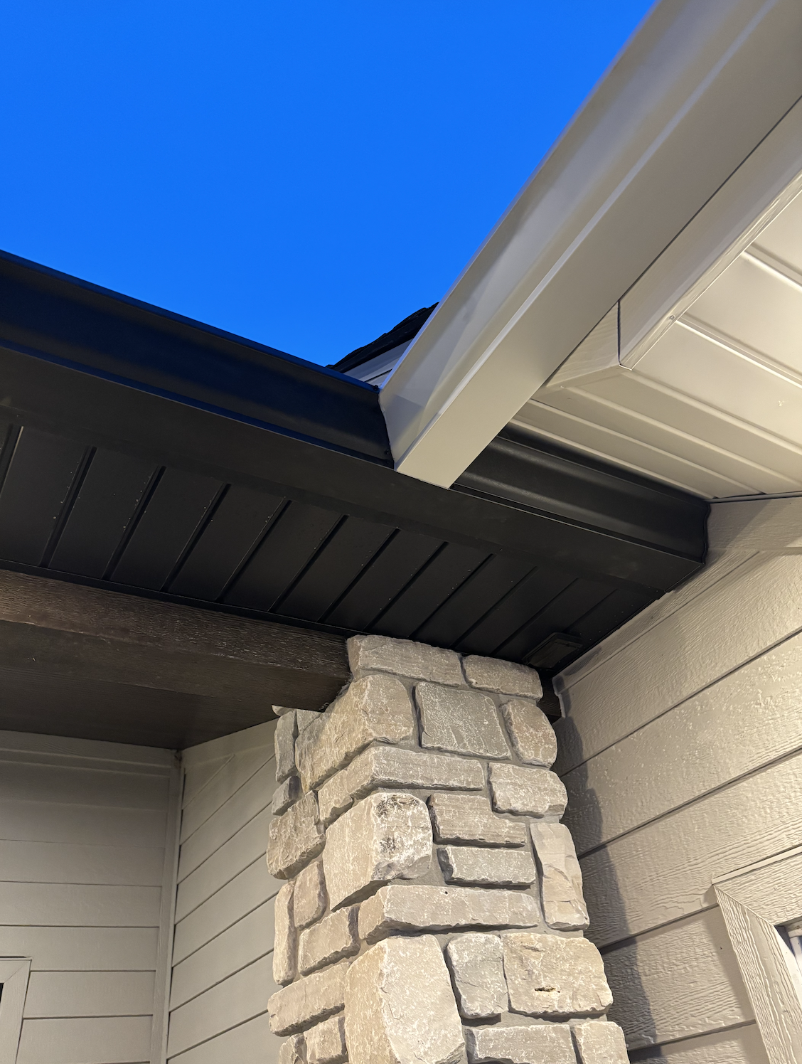 Close-up of a house corner showing beige siding, a stone pillar, and an overhanging roof with white trim and black fascia board, under a clear blue sky.