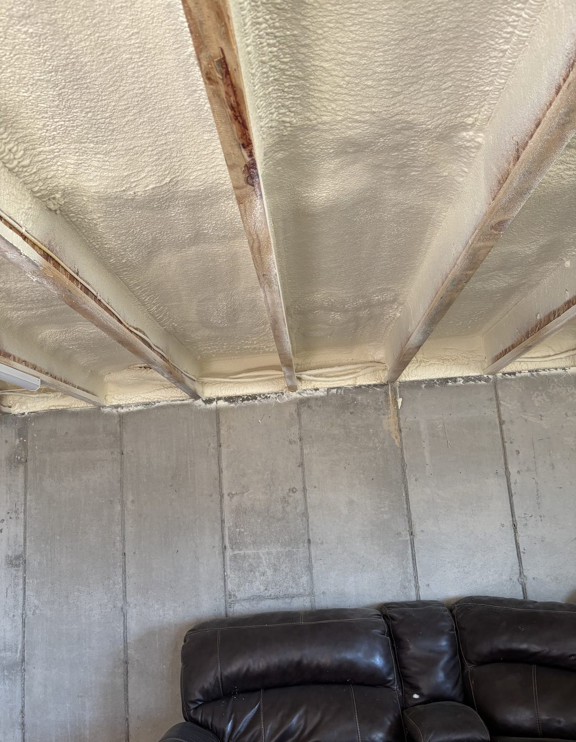 Ceiling with spray foam insulation and exposed wooden beams, unfinished wall with concrete panels, black leather couch in a basement or unfinished room.