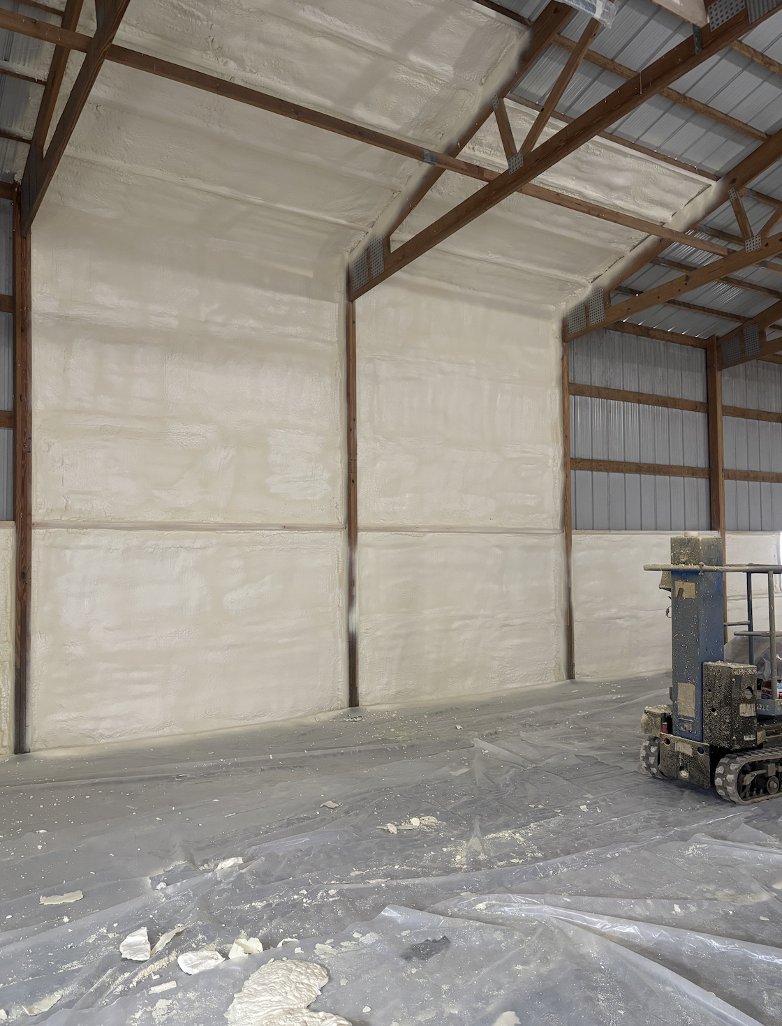 Partially constructed interior wall with spray foam insulation, a scissor lift, and a plastic covered floor