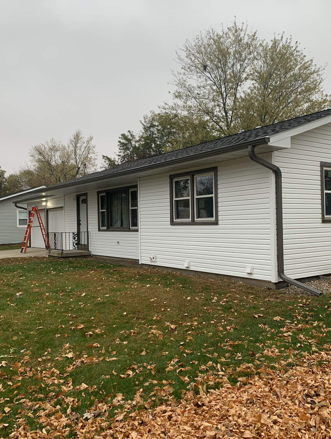 A white single-story house with black trim, three windows, a small front porch with a door, a ladder leaning against the house, and a yard covered with fallen autumn leaves.