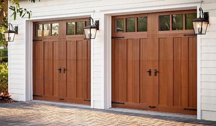 Two wooden garage doors with black hardware and small transom windows at the top, mounted on a white building with hanging lantern-style lights on each side.
