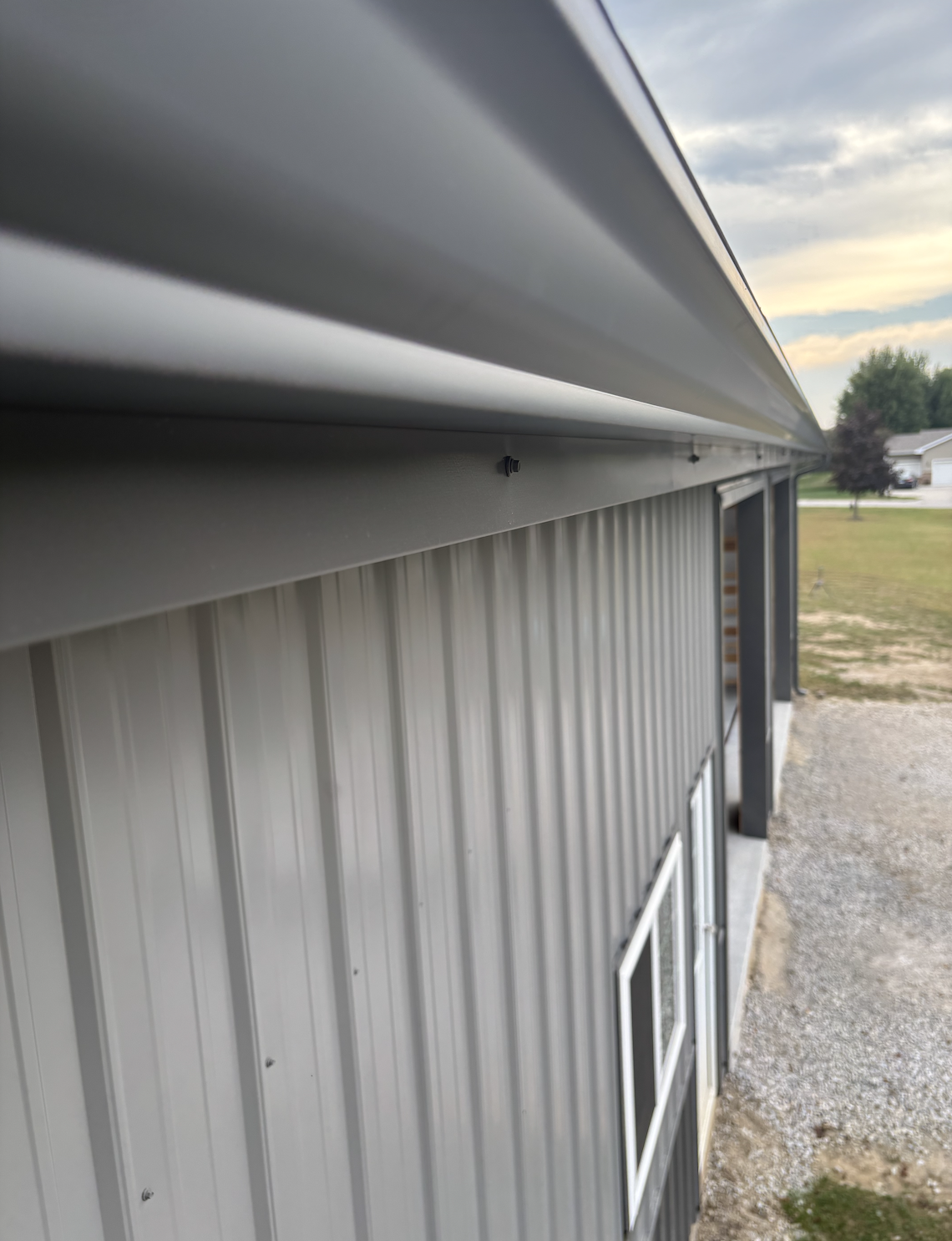 Close-up of the side of a building with metal siding and a protruding metal awning or roof, outdoors with houses, trees, and cloudy sky in the background.