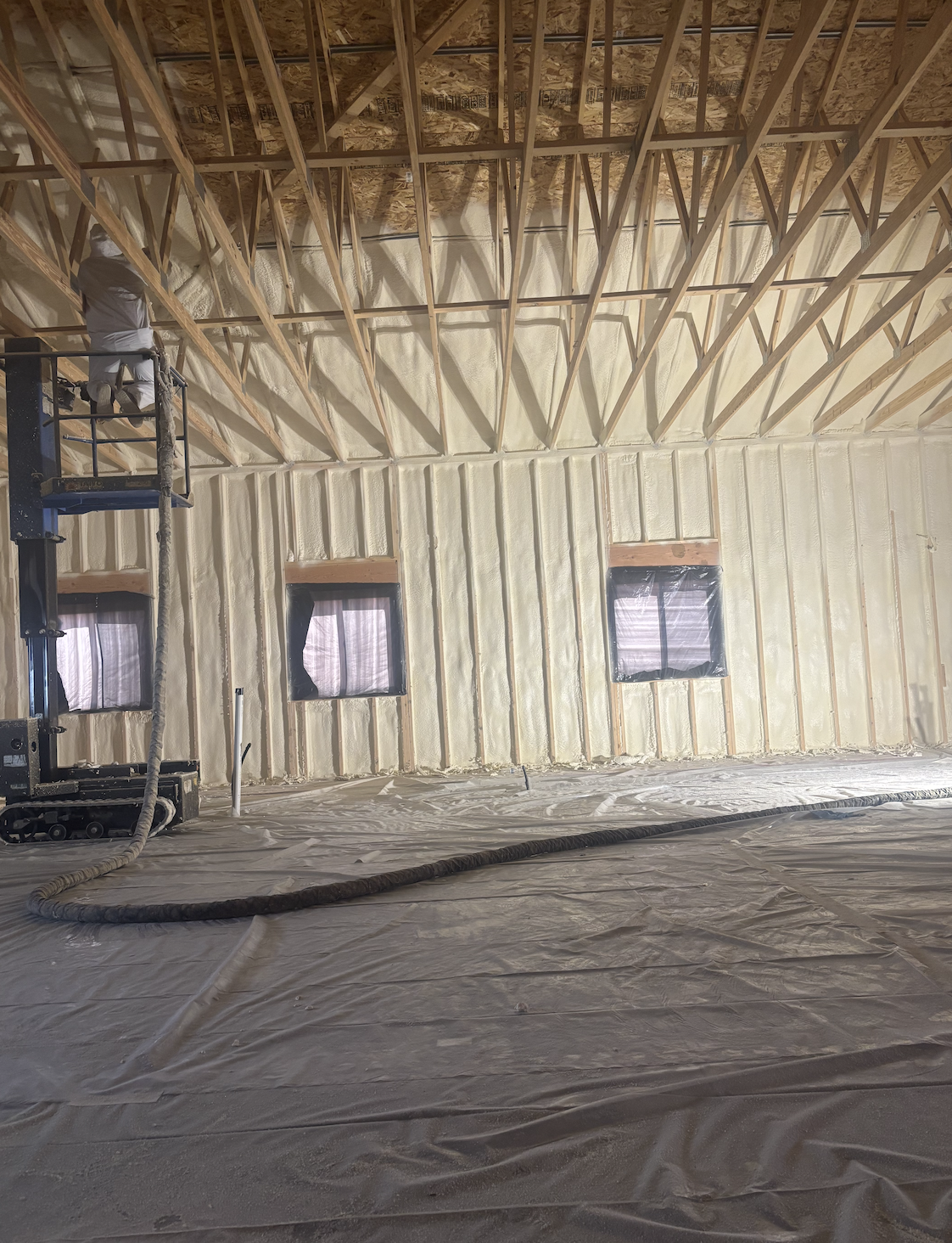 Interior of a building under construction with exposed wooden framing, spray foam insulation on the walls, and windows covered with plastic sheeting. A worker is on a lift applying insulation to the ceiling.