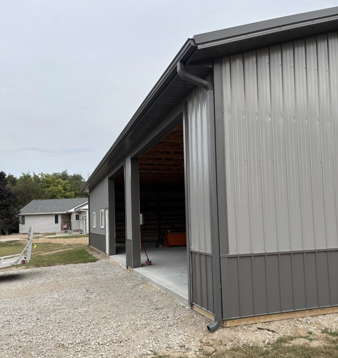A gray metal building with an open garage door, construction tools inside, gravel driveway, and neighboring houses in the background.