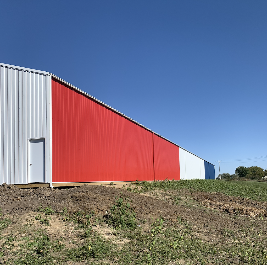 Large industrial building with red, white, and blue metal siding against a clear blue sky, with dirt and grass in the foreground.