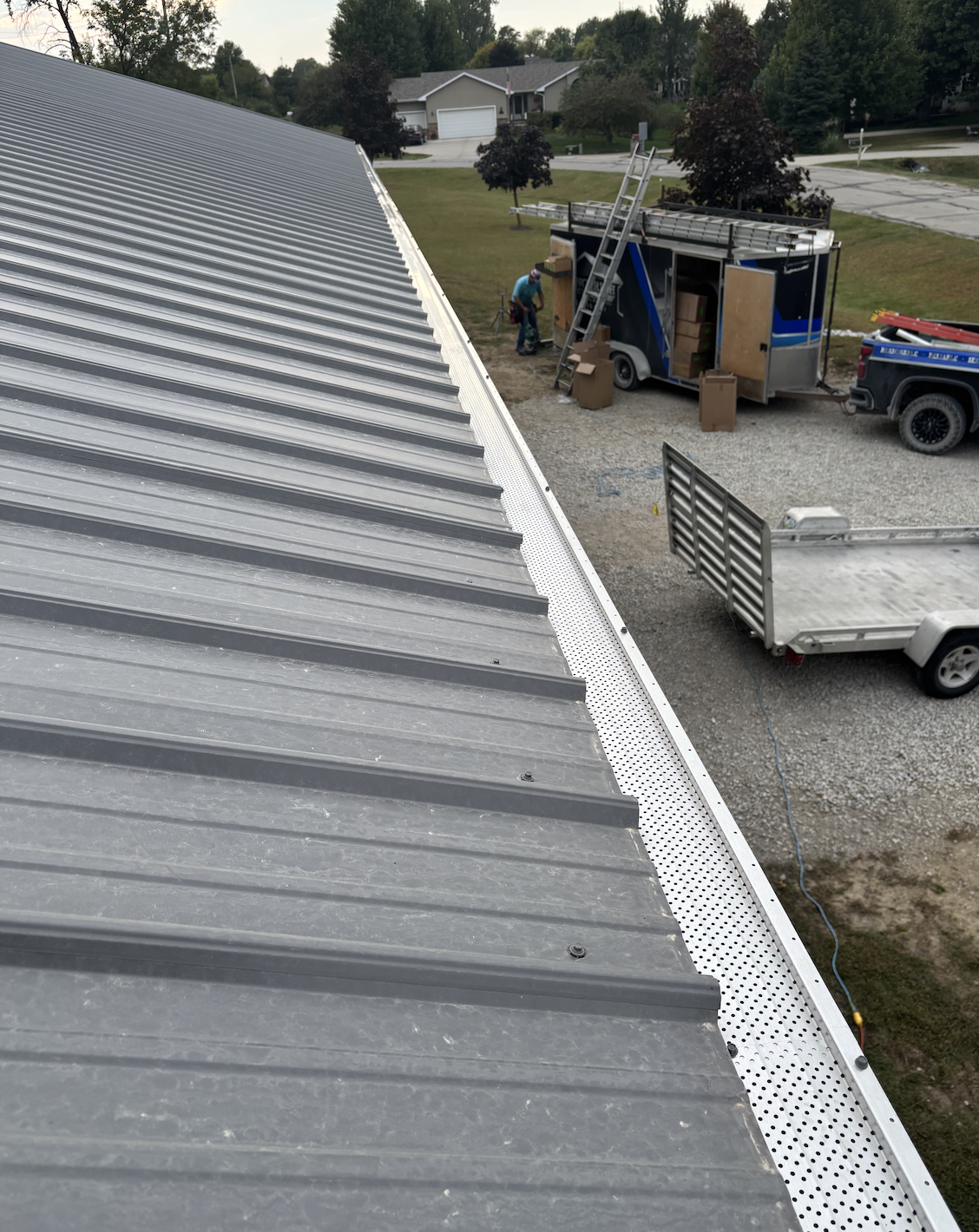 View from the roof showing a work crew loading equipment into a blue work trailer, with trees, a house, and a street in the background.