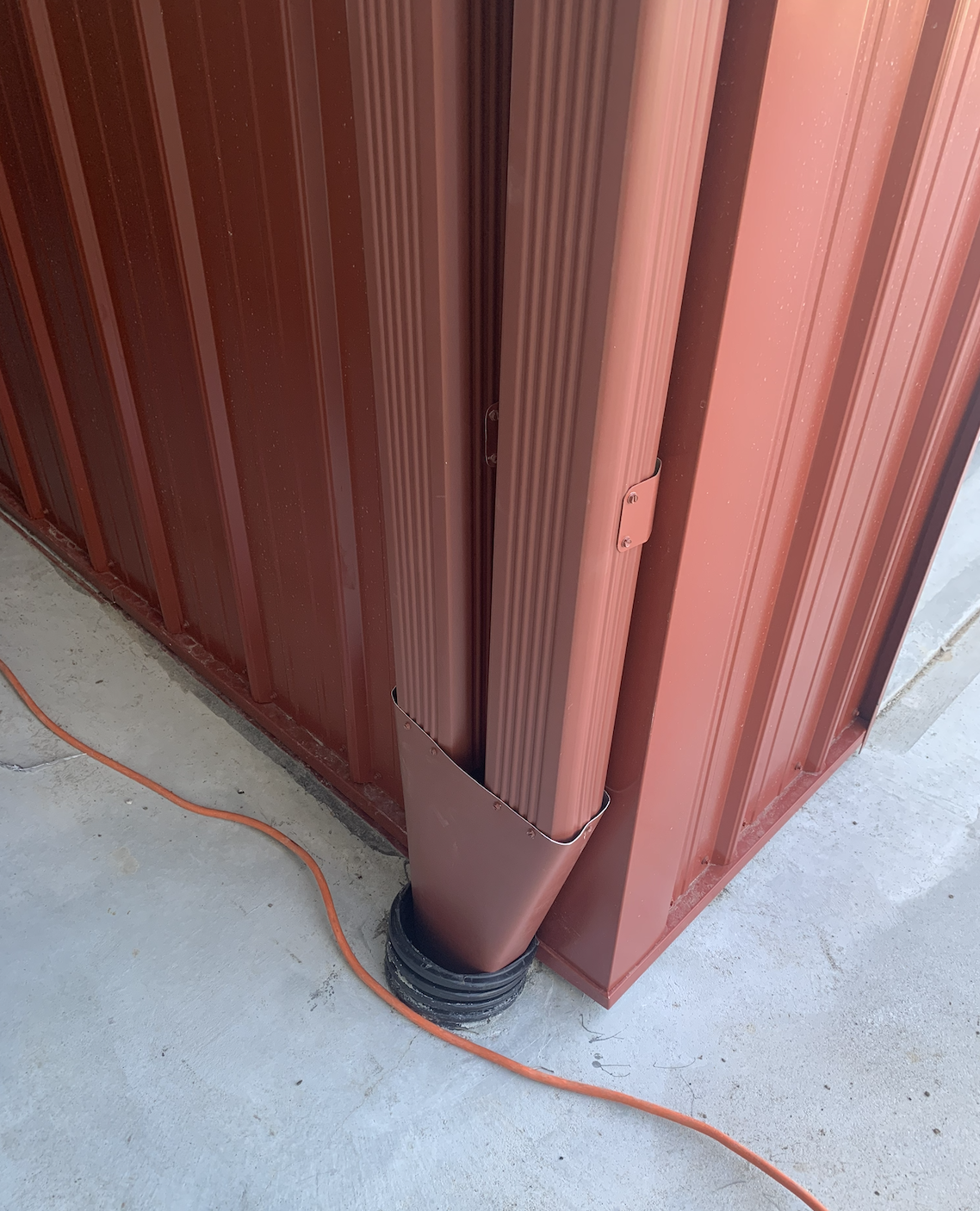 Close-up of a red metal building corner with a vertical drainage pipe and an orange extension cord on a concrete floor.