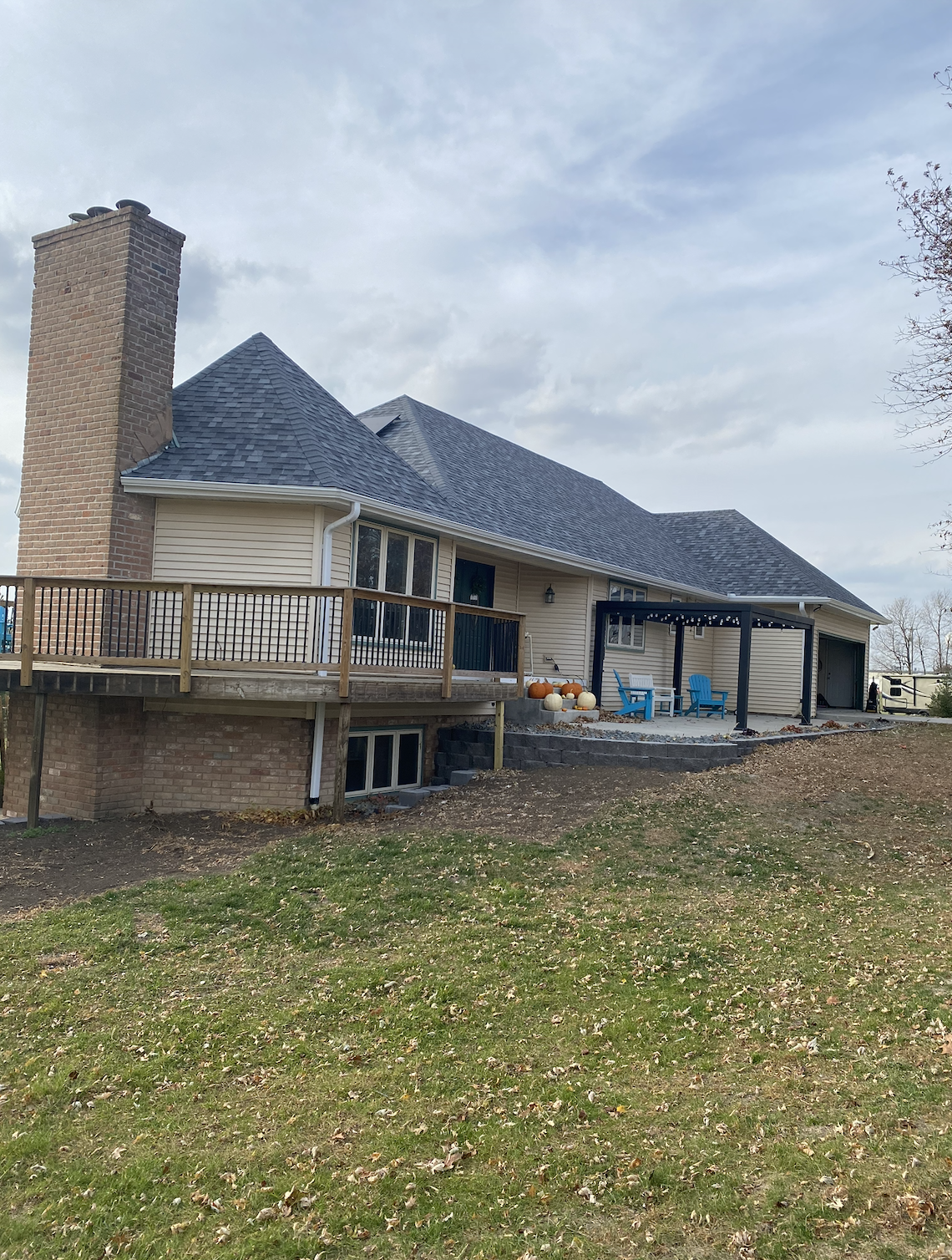 The back of a house with a brick chimney, gray sloped roof, and a wooden deck with outdoor seating and pumpkins, under a partly cloudy sky.