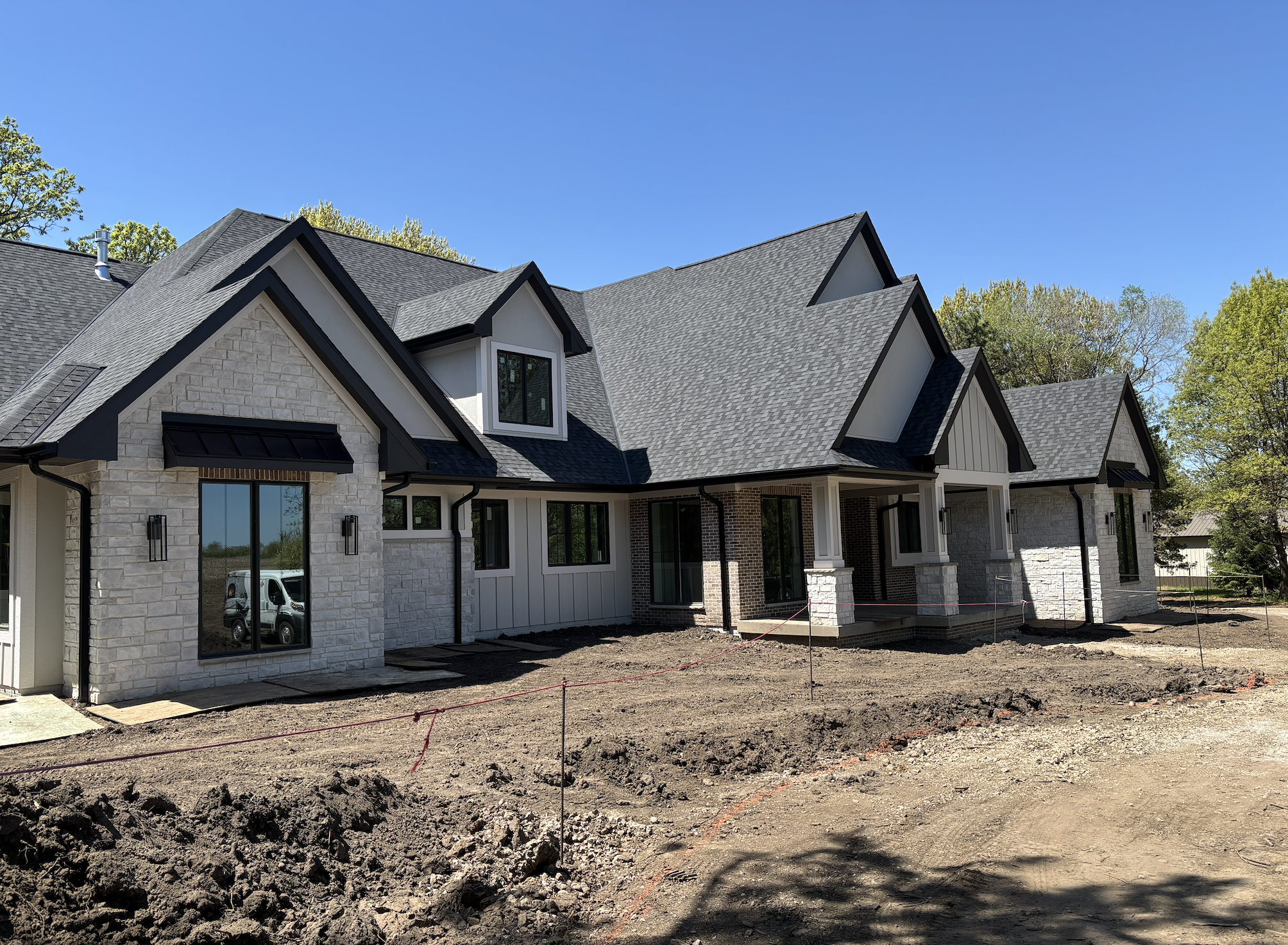 A newly built house with a steeply pitched roof, brick and white siding exterior, and multiple windows, situated on a lot under construction with visible dirt and groundwork.