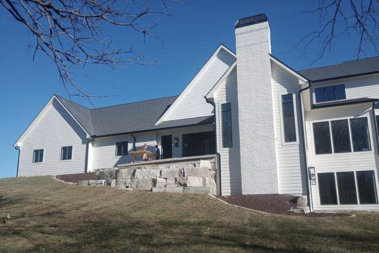 A large white house with a chimney, multiple windows, and a porch on a grassy hill under a clear blue sky, with leafless tree branches at the top of the image.