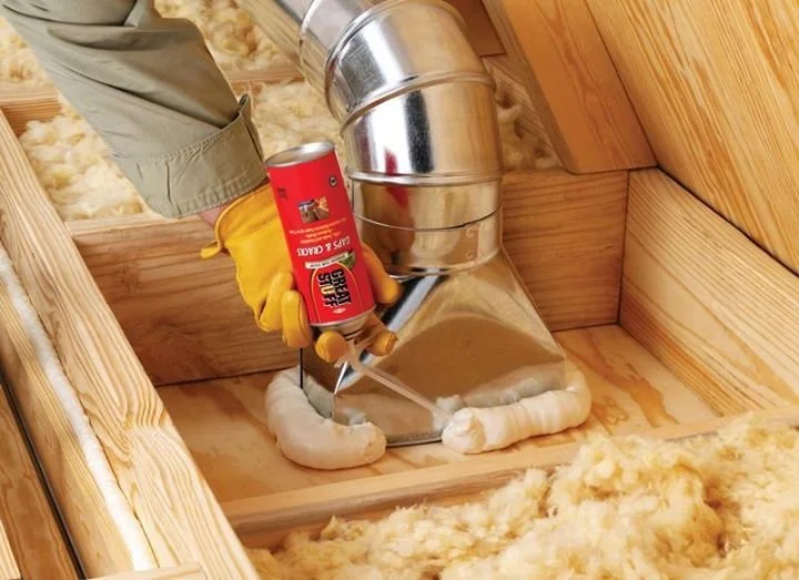 A person is installing a metal HVAC vent pipe in a wood-framed wall, using a propane torch to soften sealing foam around the vent opening, in a room with insulation and unfinished wood framing.