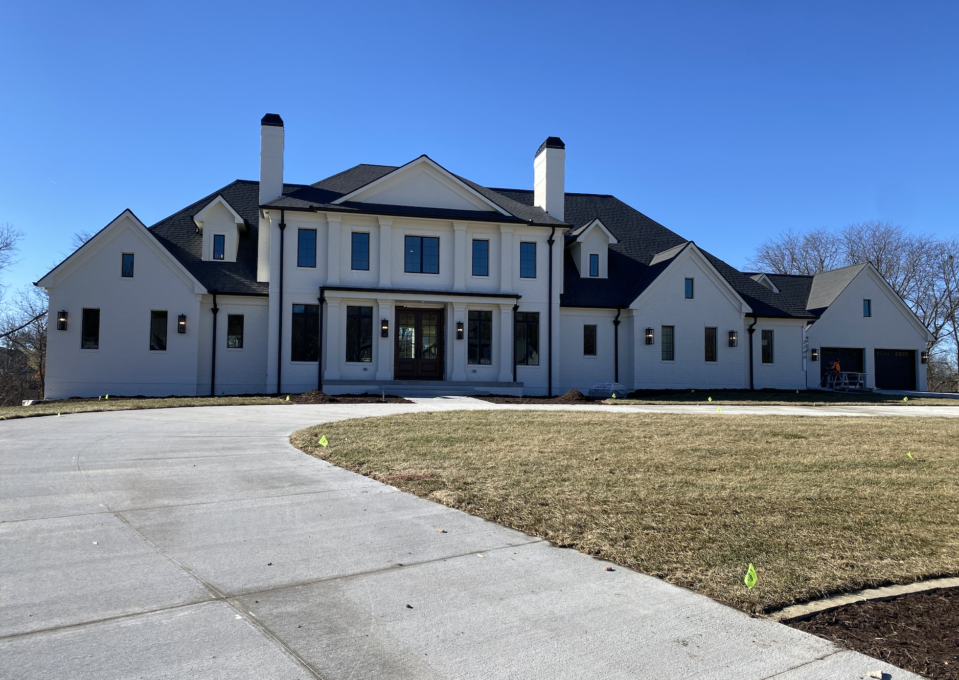 Front view of a newly built white house with black trim, multiple gables, and two prominent chimneys against a clear blue sky, with a curved driveway and a grassy lawn.