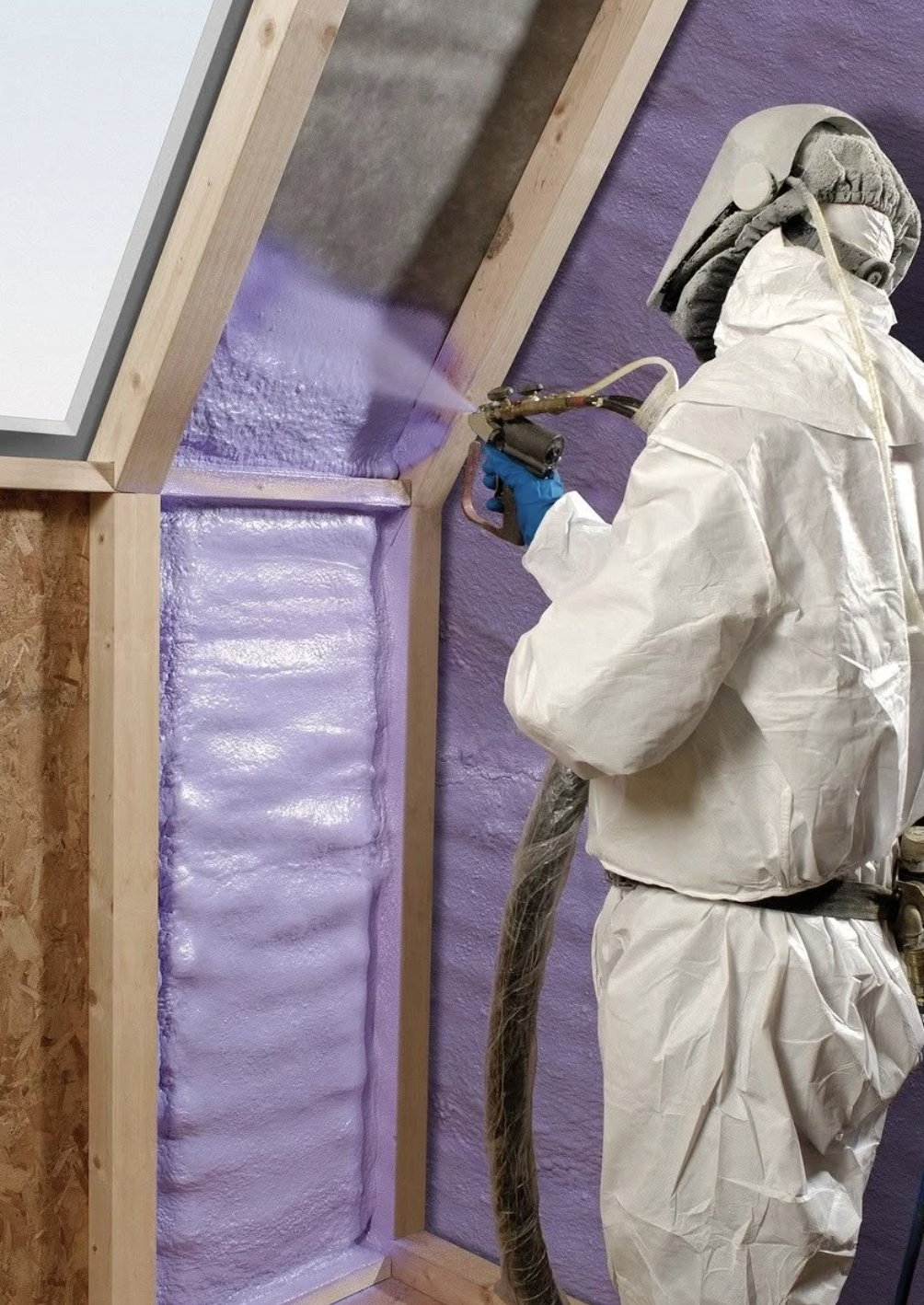 Person wearing protective suit and mask using spray foam insulation on a wall inside a building.