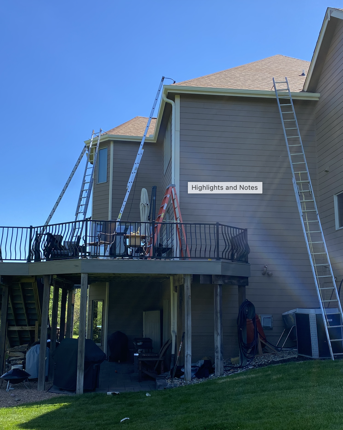 A house with outdoor deck under construction, featuring three ladders leaning against the house, patio furniture on the deck, and construction tools below.
