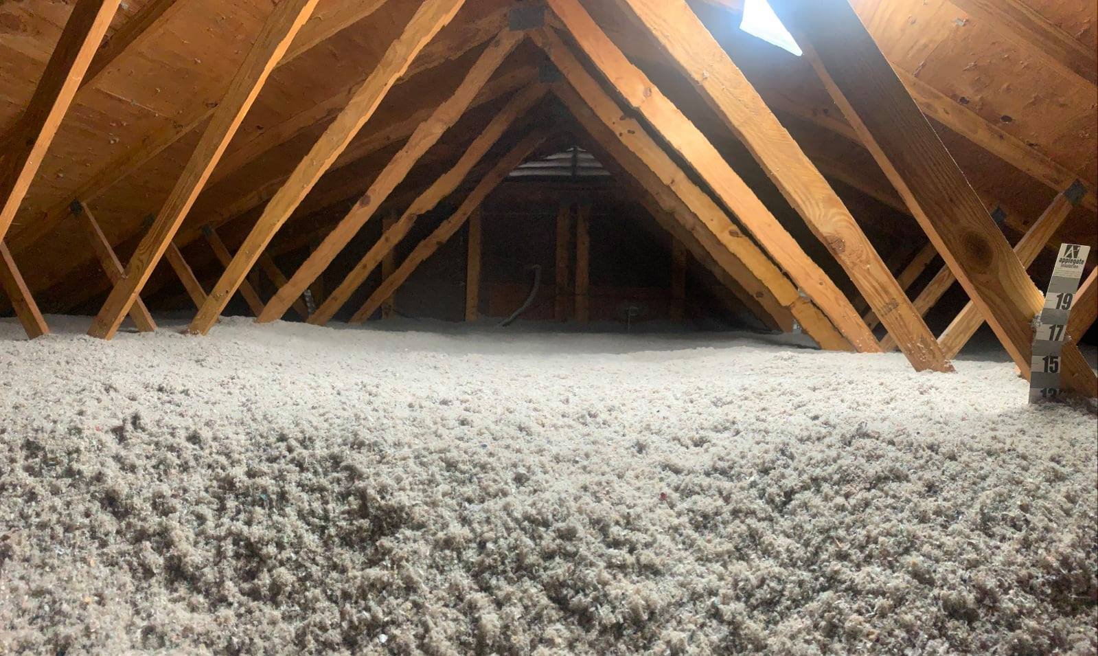Inside an attic showing wooden roof rafters, insulation material on the floor, and a height measurement scale on the right side.