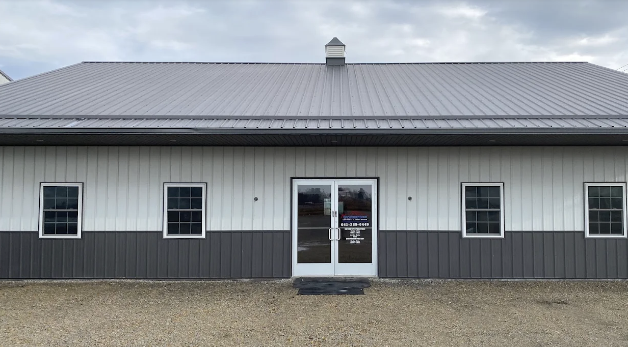 Front view of a commercial building with three windows and a glass double door, situated on a gravel lot under an overcast sky.
