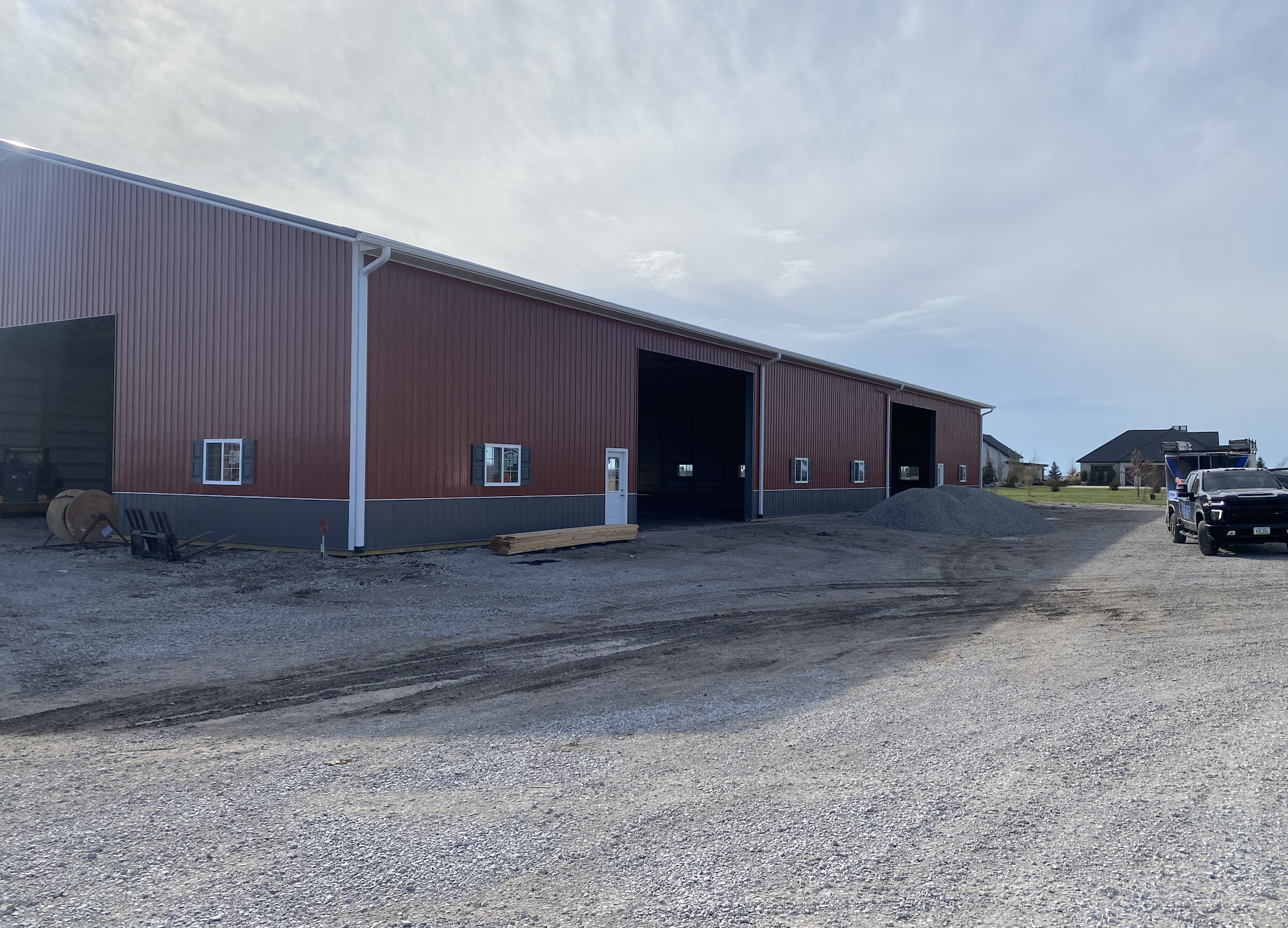 A large red metal building with open garage doors, situated in a construction area with dirt ground and construction materials. In the background, there are residential houses and a parked vehicle.