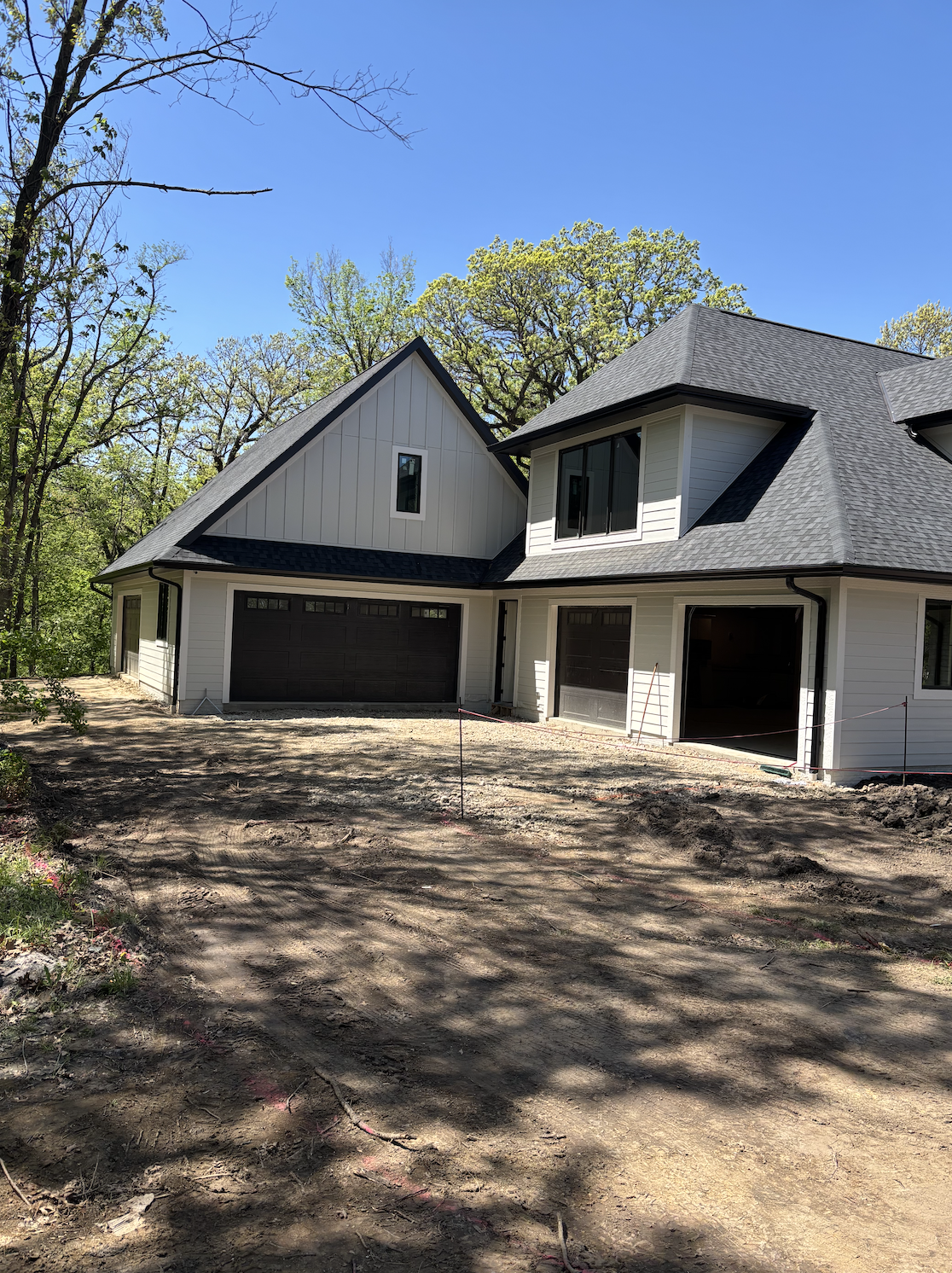 Newly constructed house with white siding and black garage doors, surrounded by trees, on a dirt lot under a clear blue sky.
