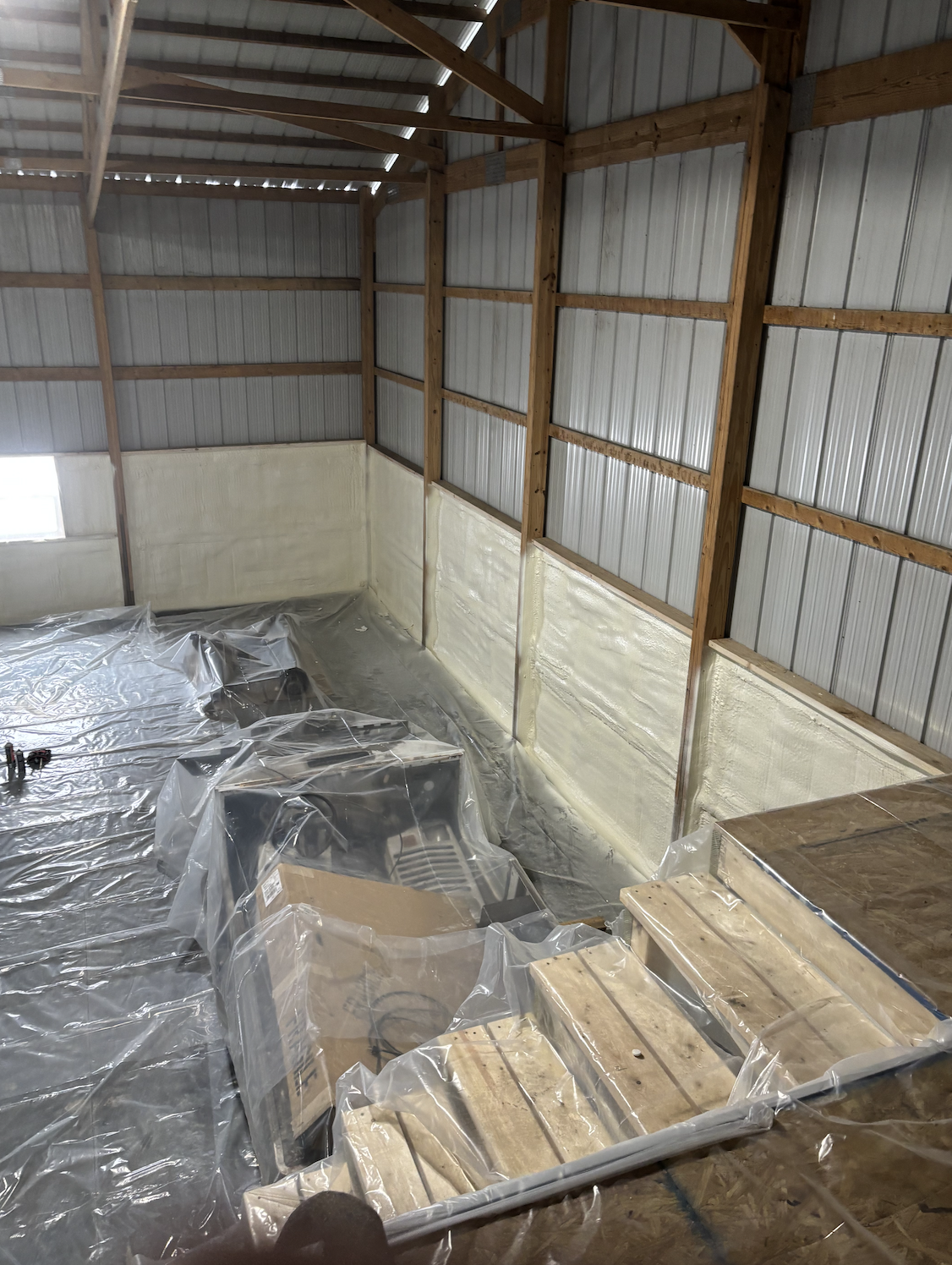 Interior of a barn or shed under construction, with wooden framing, metal siding, insulation, and plastic covering the floor and furniture.