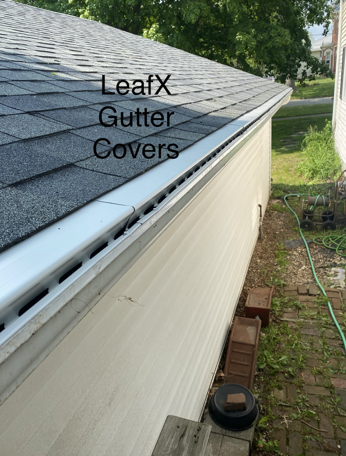 Image of a house gutter with a leaf guard cover, showing roof shingles and a side yard with garden hose and pavers.