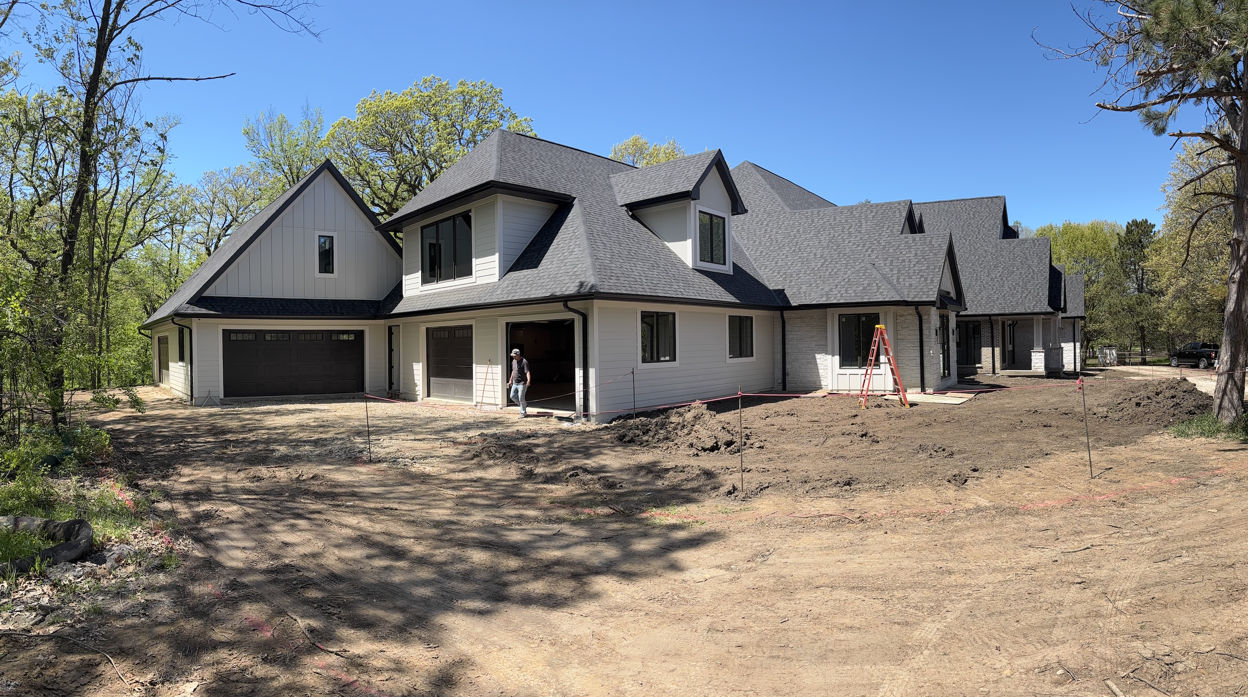 Newly built house with gray roof shingles and white siding, under construction, with a worker walking in the yard, surrounded by trees and a clear blue sky.
