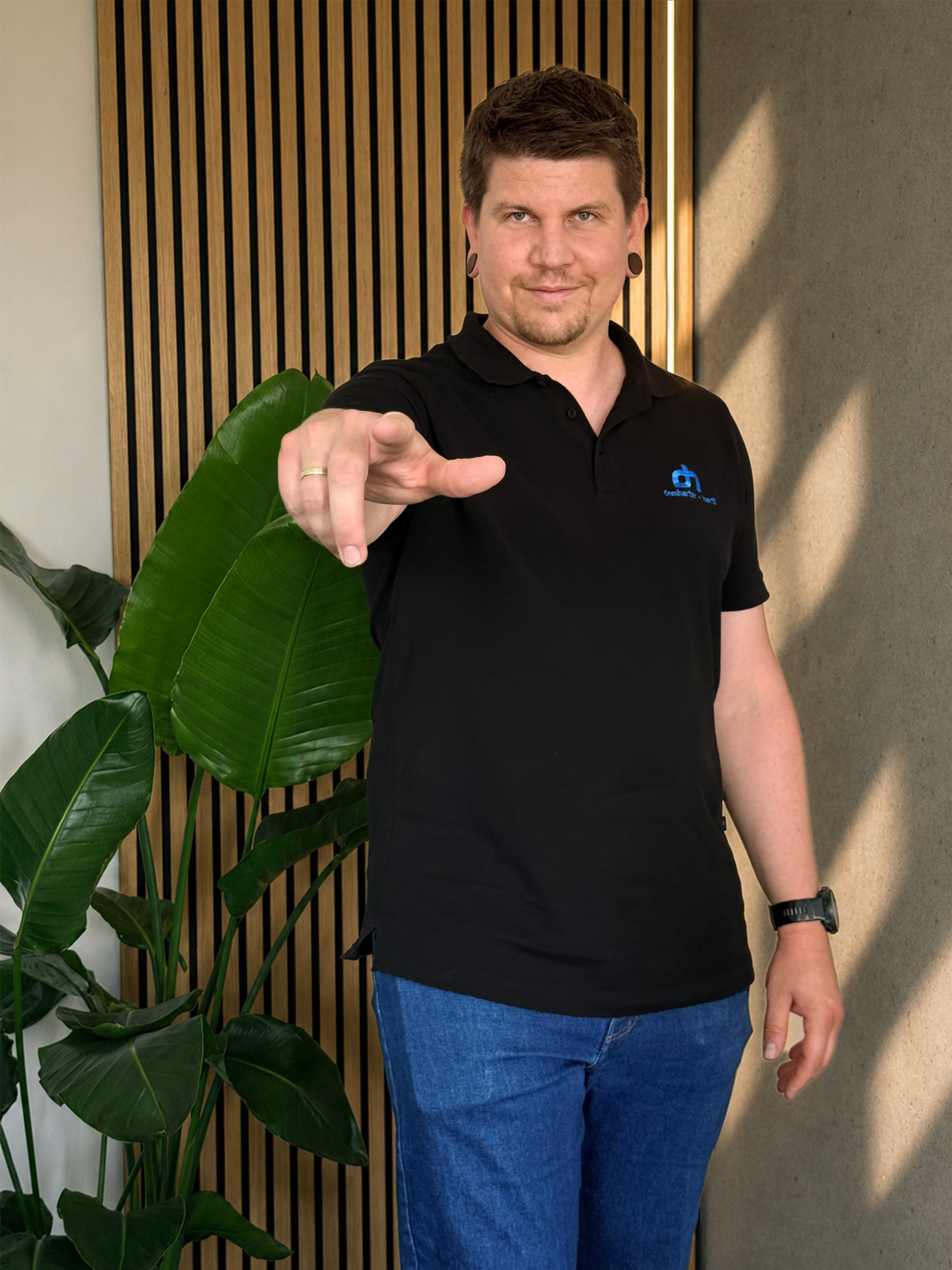 Ein Mann mit dunklem Haar, dunkelbraunen Ohrläppchen, trägt ein schwarzes Polo-Shirt mit einem blauen Logo und blaue Jeans, steht vor einer Wand mit Holzstreifen, zeigt mit der rechten Hand auf die Kamera, trägt eine schwarze Armbanduhr, und im Hintergrund sind große grüne Blätter einer Zimmerpflanze sichtbar.