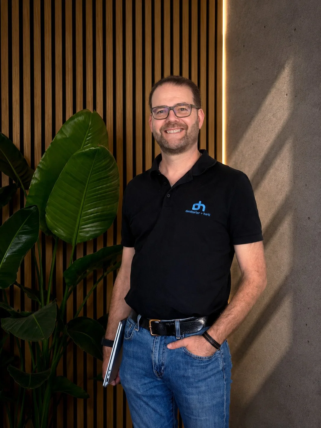 Ein Mann in schwarzem Polo mit einem blauen Logo steht vor einer Wand mit Holzelementen und einer großen Zimmerpflanze.