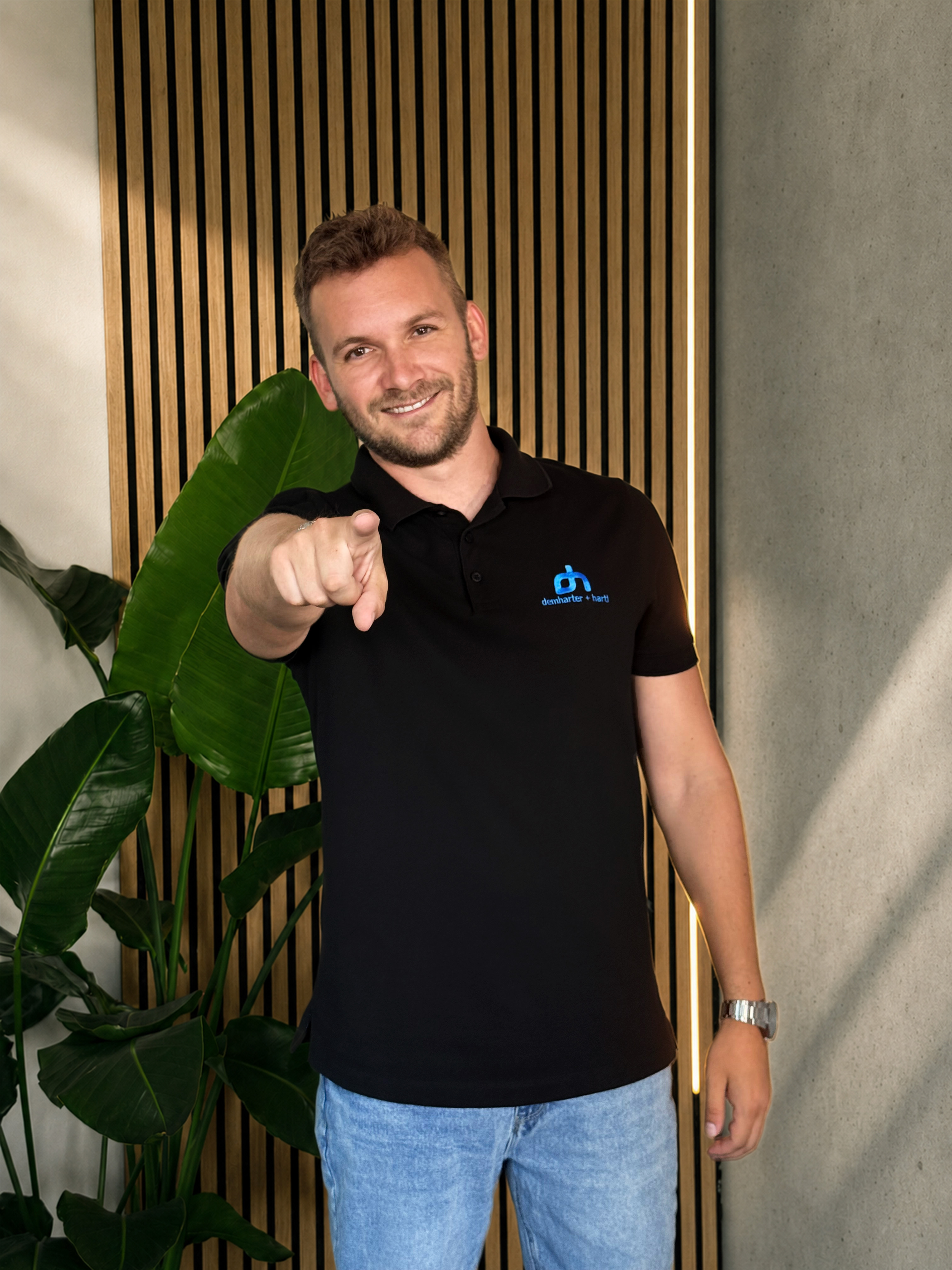 Ein Mann trägt ein schwarzes T-Shirt mit einem blauen Logo und Denim-Hosen, hält einen Arm nach vorne und zeigt auf den Betrachter, vor einer Wand mit Holzstreifen und einem großen grünen Blatt einer Pflanze.