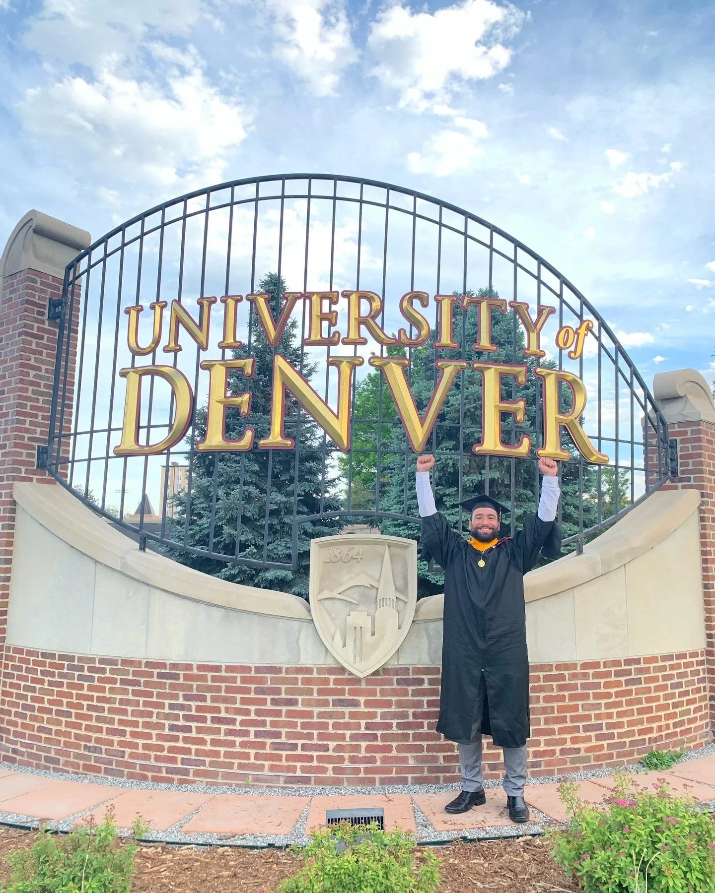 A man in a graduation gown and cap celebrating in front of the University of Denver sign, raising his fists in victory.