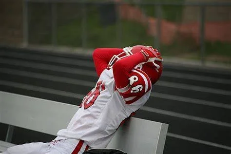 A football player in a red and white uniform sitting on a bench with his arms raised, holding his helmet.