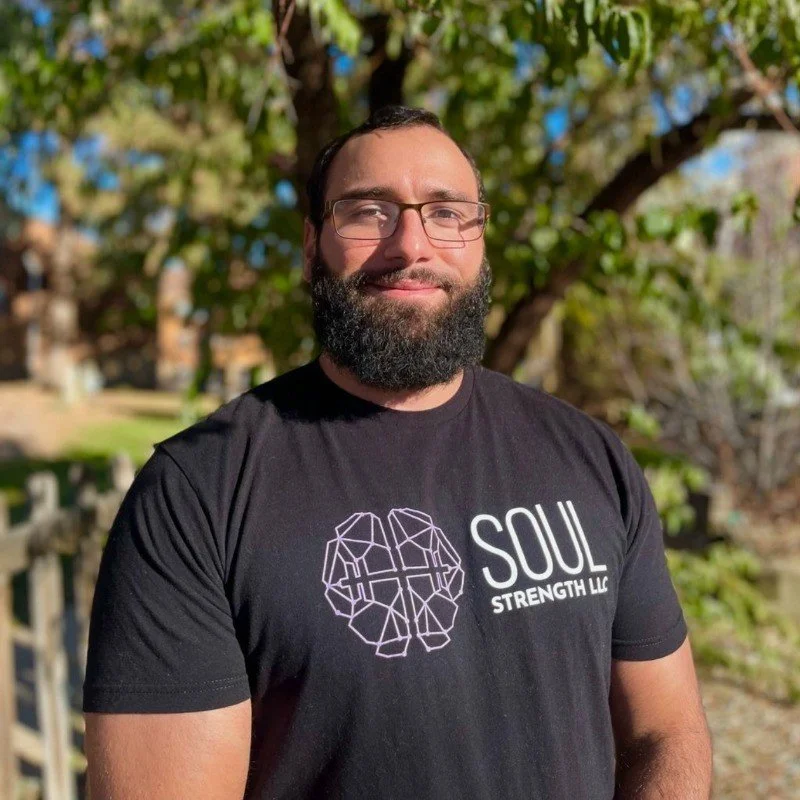A man with glasses and a beard smiling outdoors, wearing a black T-shirt with a logo and text that reads 'SOUL STRENGTH LLC', background with trees and sunlight.