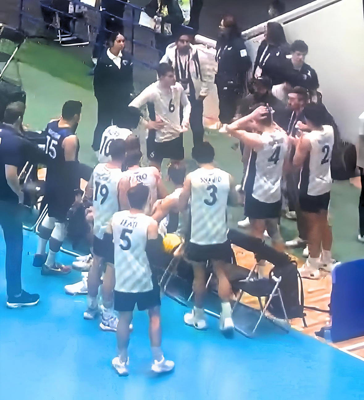 A volleyball team and staff gathered on the sidelines during a match, with some players sitting or kneeling and others standing, while discussing or receiving instructions.