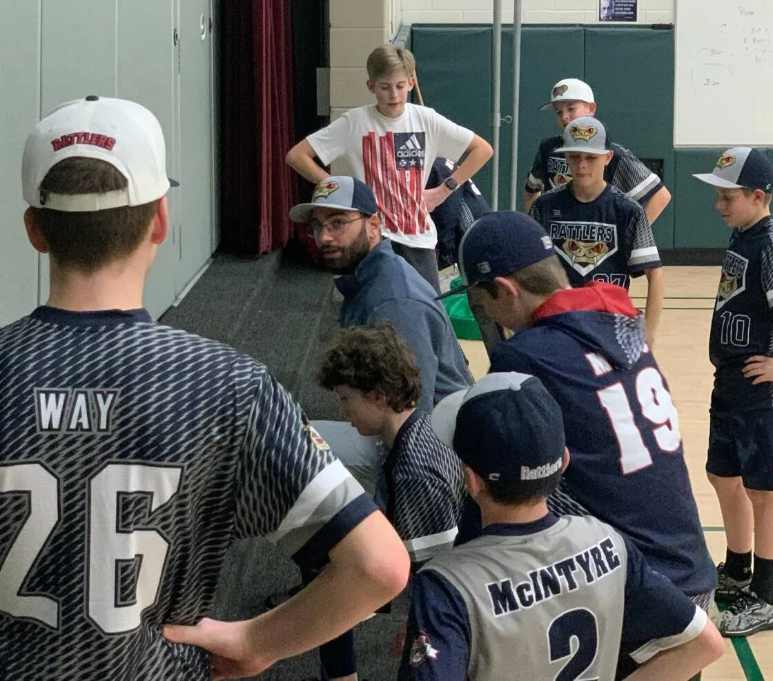 Group of young boys in sports jerseys and hats gather around a coach or adult in a gymnasium, possibly during a team meeting or discussion.