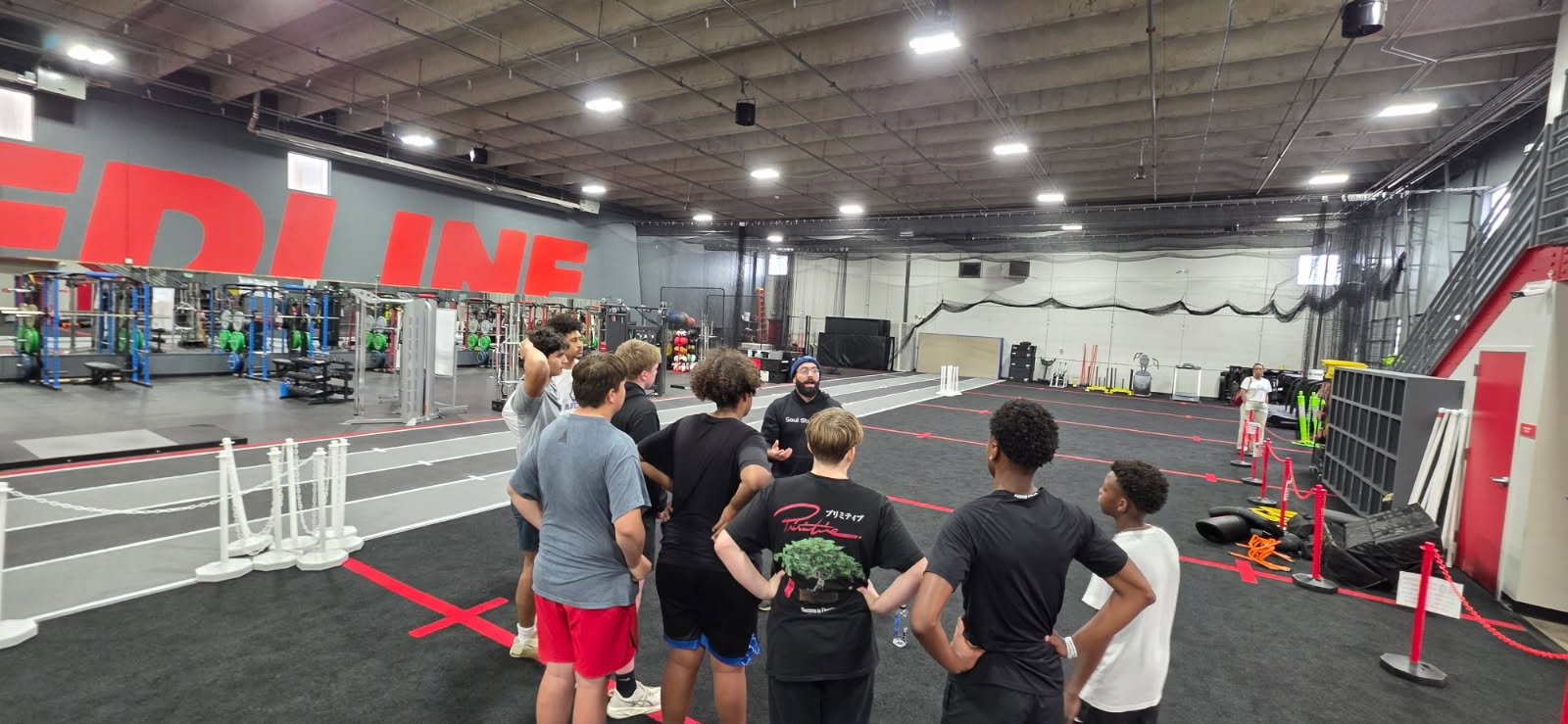 A group of young people listening to a coach in a gym with workout equipment and a running track.