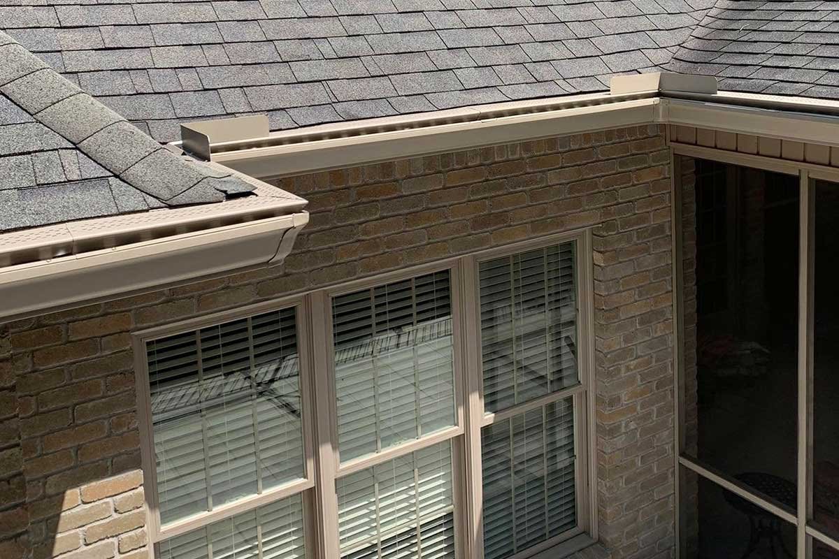 Exterior view of a brick house with two large windows and a screened-in porch on the right. The house has a gray shingled roof and a LeafX gutter protection in cream.