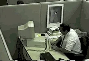 Office cubicle with a man sitting at a desk, working on a computer, with a monitor, stacks of papers, and a framed photo on the wall.