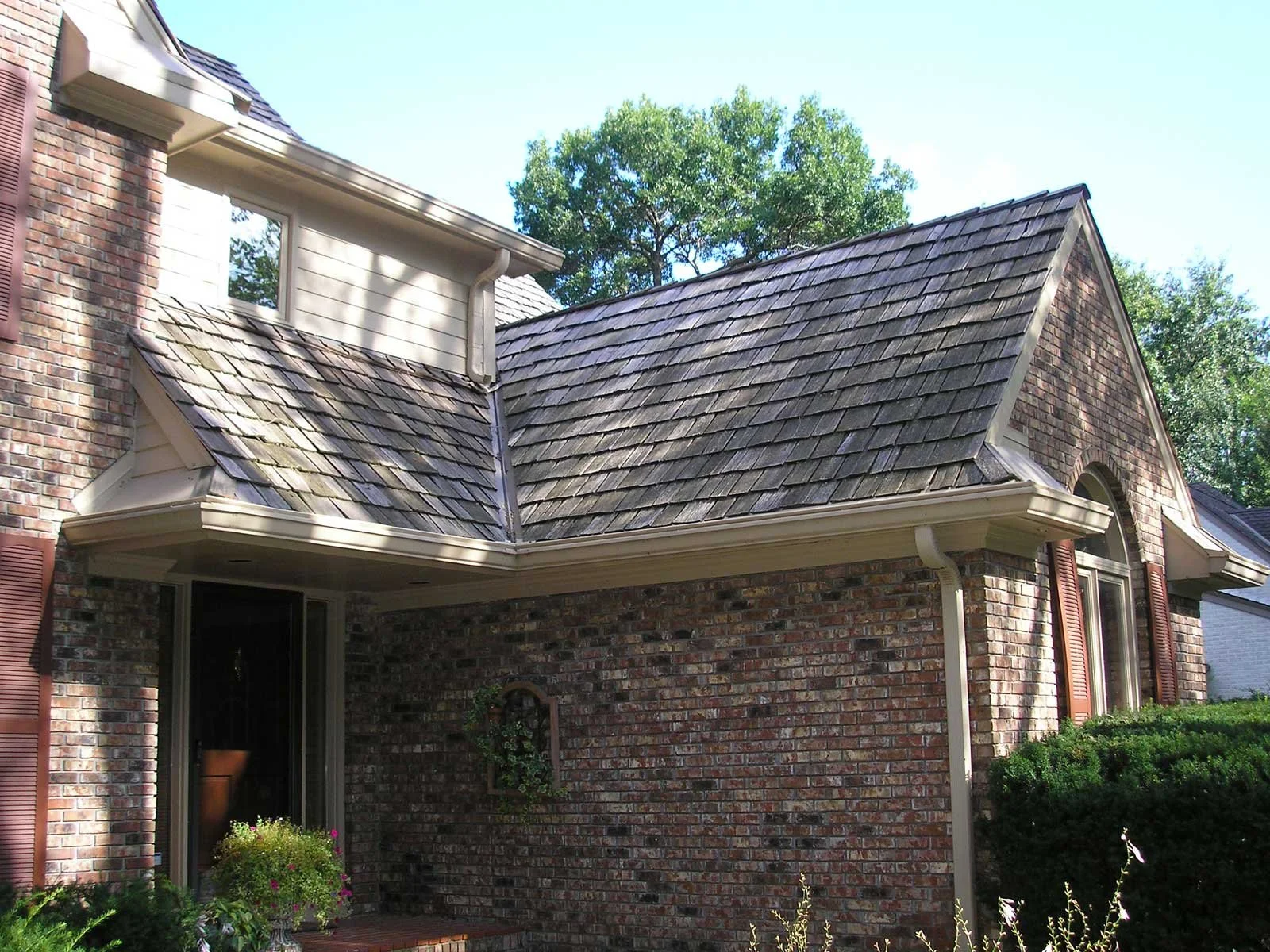 A brick house with a gabled roof, shingle siding, and a small porch area. There are trees in the background and some plants in front of the house.