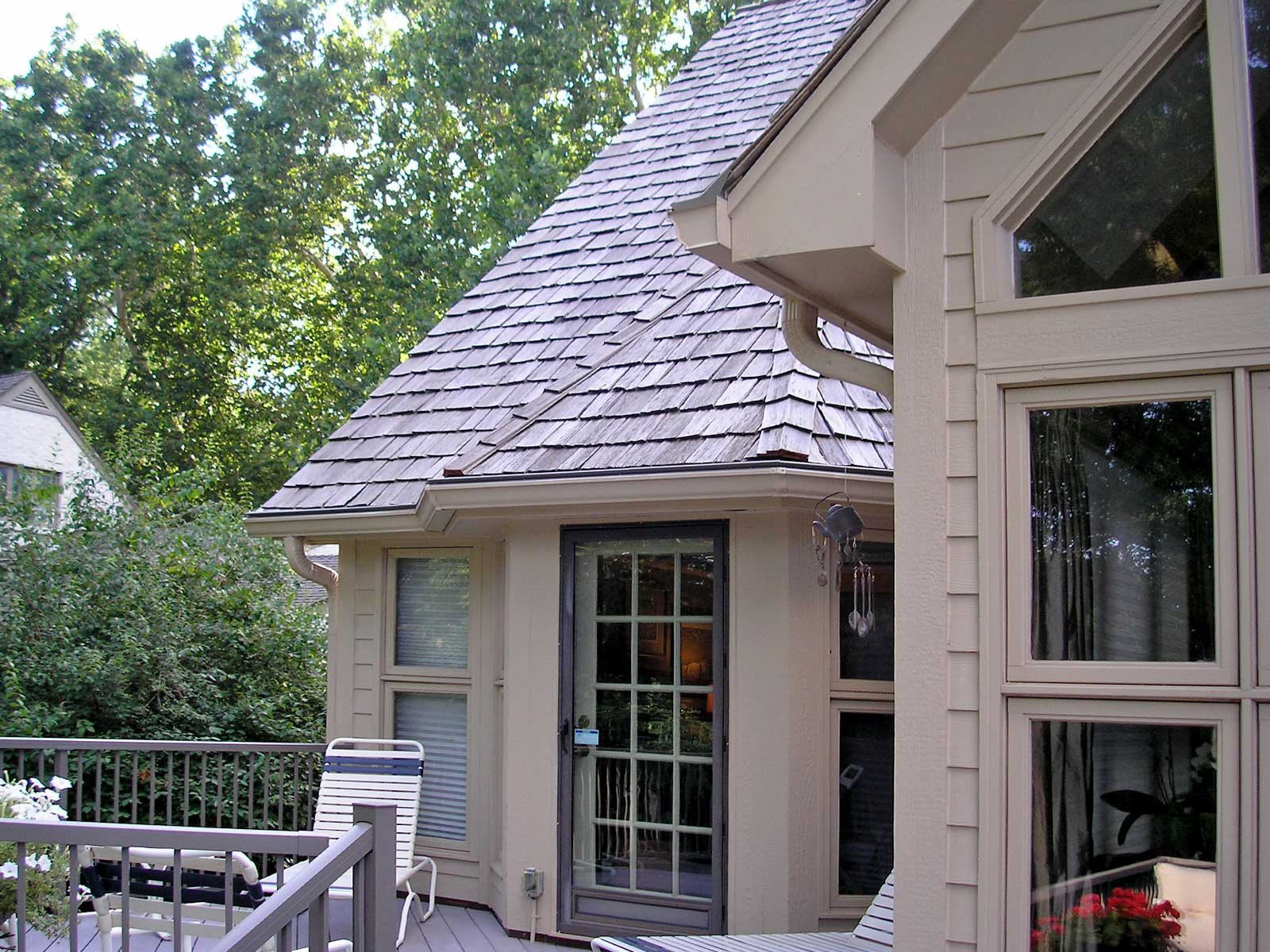 Backyard patio with beige house and a deck, featuring a door, large windows, outdoor chairs, and surrounded by green trees.